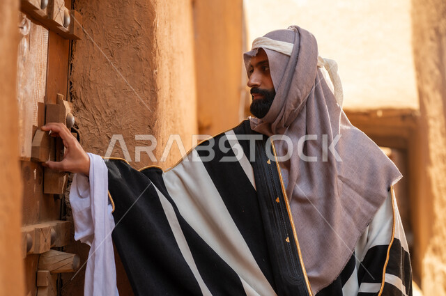 رجل عربي سعودي خليجي يرتدي زي يوم التأسيسي يقف ويمسك باب مبنى طيني، ذكرى تأسيس الدوله السعوديه الاولى على يد الامام محمد بن سعود, يوم بدينا 22 فبراير 1727م, العمق التاريخي والحضاري للمملكة، هوية اليوم الوطني السعودي 92
