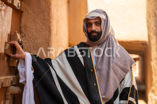رجل عربي سعودي خليجي يرتدي زي يوم التأسيسي يقف ويمسك باب مبنى طيني، ذكرى تأسيس الدوله السعوديه الاولى على يد الامام محمد بن سعود, يوم بدينا 22 فبراير 1727م, العمق التاريخي والحضاري للمملكة، هوية اليوم الوطني السعودي 92