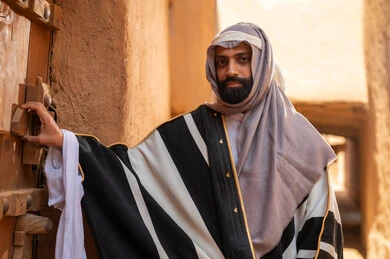 A Saudi Arabian Gulf man wearing the founding day attire stands holding the door of a mud building, commemorating the establishment of the first Saudi state by Imam Muhammad bin Saud, on the day we began, February 22, 1727 AD, the historical and cultural depth of the Kingdom, the identity of Saudi National Day 92.