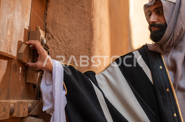 رجل عربي سعودي خليجي يرتدي زي يوم التأسيسي يقف ويمسك باب مبنى طيني، ذكرى تأسيس الدوله السعوديه الاولى على يد الامام محمد بن سعود, يوم بدينا 22 فبراير 1727م, العمق التاريخي والحضاري للمملكة، هوية اليوم الوطني السعودي 92