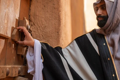 رجل عربي سعودي خليجي يرتدي زي يوم التأسيسي يقف ويمسك باب مبنى طيني، ذكرى تأسيس الدوله السعوديه الاولى على يد الامام محمد بن سعود, يوم بدينا 22 فبراير 1727م, العمق التاريخي والحضاري للمملكة، هوية اليوم الوطني السعودي 92