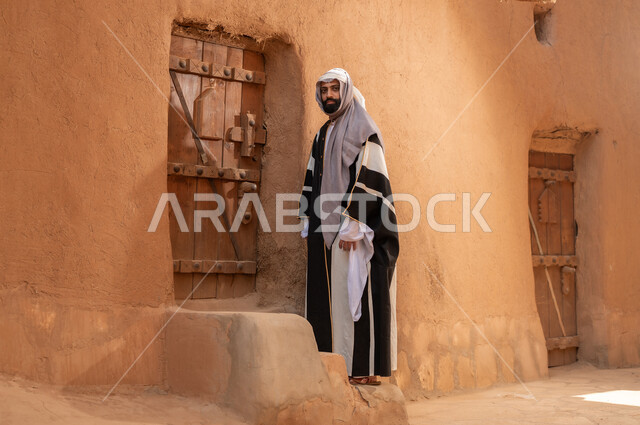 رجل عربي سعودي خليجي يرتدي زي يوم التأسيسي يقف بجانب مبنى طيني، ذكرى تأسيس الدوله السعوديه الاولى على يد الامام محمد بن سعود, يوم بدينا 22 فبراير 1727م, العمق التاريخي والحضاري للمملكة، هوية اليوم الوطني السعودي 92