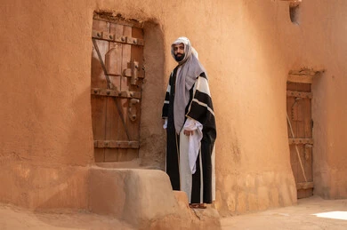 A Saudi Arabian Gulf man wearing the founding day attire stands next to a mud building, commemorating the establishment of the first Saudi state by Imam Muhammad bin Saud, on the day we began, February 22, 1727 AD, the historical and cultural depth of the Kingdom, the identity of Saudi National Day 92.