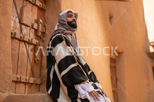صورة من الجانب رجل عربي سعودي خليجي يرتدي زي يوم التأسيسي يجلس على سلم مبنى طيني، ذكرى تأسيس الدوله السعوديه الاولى على يد الامام محمد بن سعود, يوم بدينا 22 فبراير 1727م, العمق التاريخي والحضاري للمملكة، هوية اليوم الوطني السعودي 92