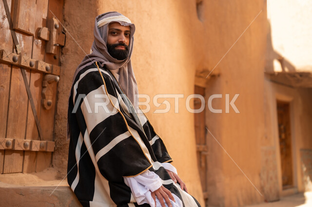 صورة من الجانب رجل عربي سعودي خليجي يرتدي زي يوم التأسيسي يجلس على سلم مبنى طيني، ذكرى تأسيس الدوله السعوديه الاولى على يد الامام محمد بن سعود, يوم بدينا 22 فبراير 1727م, العمق التاريخي والحضاري للمملكة، هوية اليوم الوطني السعودي 92