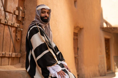 صورة من الجانب رجل عربي سعودي خليجي يرتدي زي يوم التأسيسي يجلس على سلم مبنى طيني، ذكرى تأسيس الدوله السعوديه الاولى على يد الامام محمد بن سعود, يوم بدينا 22 فبراير 1727م, العمق التاريخي والحضاري للمملكة، هوية اليوم الوطني السعودي 92