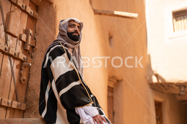 A side view of an Arab Saudi Gulf man wearing the founding day attire sitting on the steps of a clay building, commemorating the establishment of the first Saudi state by Imam Muhammad bin Saud, on the day we began, February 22, 1727 AD, the historical and cultural depth of the Kingdom, the identity of Saudi National Day 92.