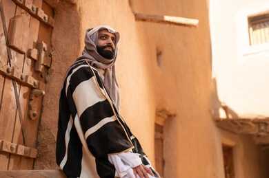 صورة من الجانب رجل عربي سعودي خليجي يرتدي زي يوم التأسيسي يجلس على سلم مبنى طيني، ذكرى تأسيس الدوله السعوديه الاولى على يد الامام محمد بن سعود, يوم بدينا 22 فبراير 1727م, العمق التاريخي والحضاري للمملكة، هوية اليوم الوطني السعودي 92