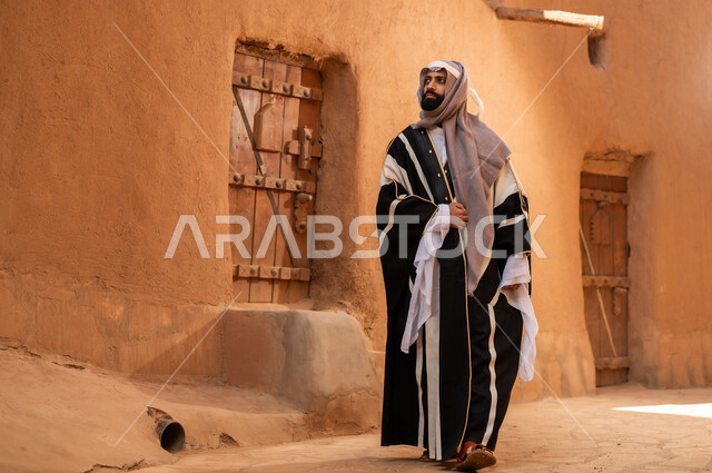 رجل عربي سعودي خليجي يرتدي زي يوم التأسيسي يقف بجانب مبنى طيني، ذكرى تأسيس الدوله السعوديه الاولى على يد الامام محمد بن سعود, يوم بدينا 22 فبراير 1727م, العمق التاريخي والحضاري للمملكة، هوية اليوم الوطني السعودي 92