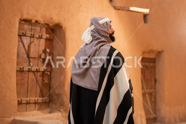 صورة من الخلف رجل عربي سعودي خليجي يرتدي زي يوم التأسيس يقف بجانب مبنى طيني, ذكرى تأسيس الدوله السعوديه الاولى على يد الامام محمد بن سعود, يوم بدينا 22 فبراير 1727م, العمق التاريخي والحضاري للمملكة، هوية اليوم الوطني السعودي 92