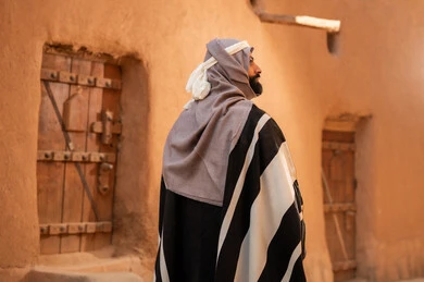 A picture from the back of an Arab Saudi Gulf man wearing the Foundation Day attire standing next to a mud building, commemorating the establishment of the first Saudi state by Imam Muhammad bin Saud, the day we began on February 22, 1727, the historical and cultural depth of the Kingdom, the identity of Saudi National Day 92.