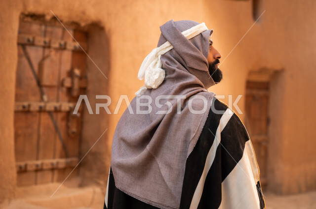 صورة من الخلف رجل عربي سعودي خليجي يرتدي زي يوم التأسيس يقف بجانب مبنى طيني, ذكرى تأسيس الدوله السعوديه الاولى على يد الامام محمد بن سعود, يوم بدينا 22 فبراير 1727م, العمق التاريخي والحضاري للمملكة، هوية اليوم الوطني السعودي 92