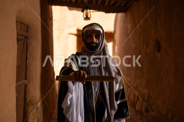 صورة من الجانب رجل عربي سعودي خليجي يرتدي زي يوم التأسيسي يقف داخل  مبنى طيني يحمل لفافة ورقية, ذكرى تأسيس الدوله السعوديه الاولى على يد الامام محمد بن سعود, يوم بدينا 22 فبراير 1727م, العمق التاريخي والحضاري للمملكة، هوية اليوم الوطني السعودي 92