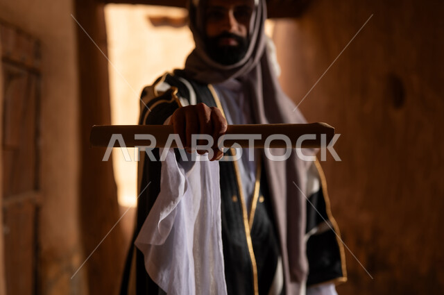 A side view of an Arab Saudi Gulf man wearing the founding day attire standing inside a clay building holding a paper scroll, commemorating the establishment of the first Saudi state by Imam Muhammad bin Saud, on the day we began, February 22, 1727 AD, the historical and cultural depth of the Kingdom, the identity of Saudi National Day 92.
