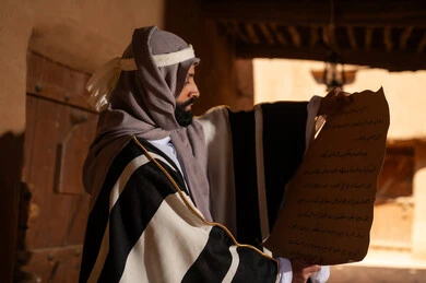 A side view of an Arab Saudi Gulf man wearing the founding day attire, standing inside and opening a paper scroll of a clay building, commemorating the establishment of the first Saudi state by Imam Muhammad bin Saud, the day we began on February 22, 1727, the historical and cultural depth of the Kingdom, the identity of Saudi National Day 92.