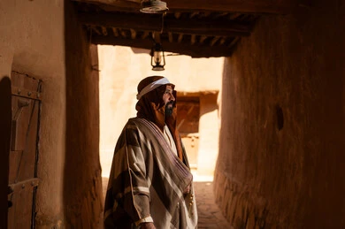 صورة من الجانب رجل عربي سعودي خليجي يرتدي زي يوم التأسيسي يقف داخل مبنى طيني, ذكرى تأسيس الدوله السعوديه الاولى على يد الامام محمد بن سعود, يوم بدينا 22 فبراير 1727م, العمق التاريخي والحضاري للمملكة، هوية اليوم الوطني السعودي 92