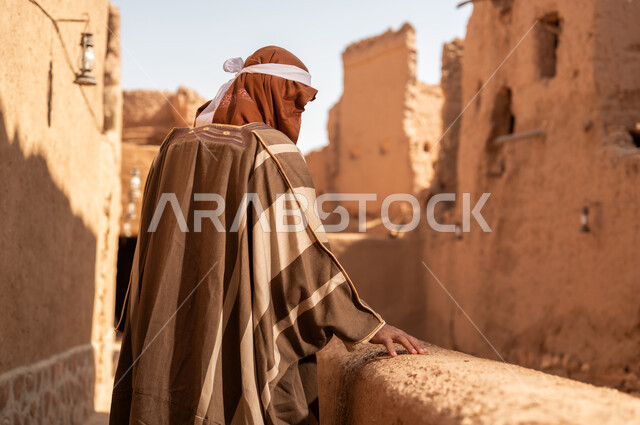 A picture from the back of an Arab Saudi Gulf man wearing the Foundation Day attire standing next to a mud building, commemorating the establishment of the first Saudi state by Imam Muhammad bin Saud, the day we began on February 22, 1727, the historical and cultural depth of the Kingdom, the identity of Saudi National Day 92.
