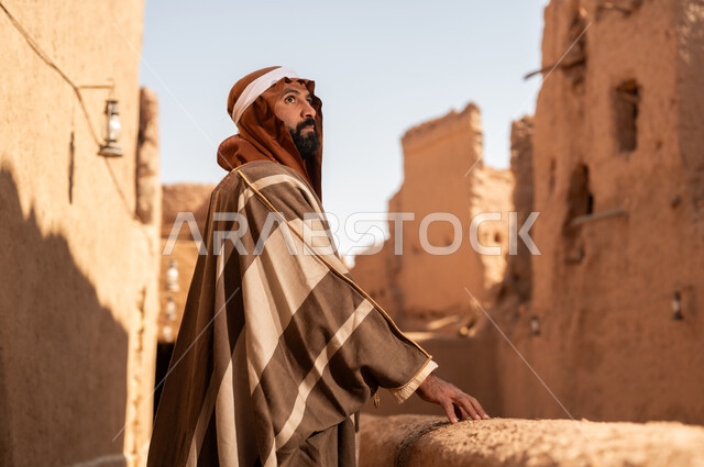 صورة من الجانب رجل عربي سعودي خليجي يرتدي زي يوم التأسيس يقف بجانب مبنى طيني, ذكرى تأسيس الدوله السعوديه الاولى على يد الامام محمد بن سعود, يوم بدينا 22 فبراير 1727م, العمق التاريخي والحضاري للمملكة، هوية اليوم الوطني السعودي 92