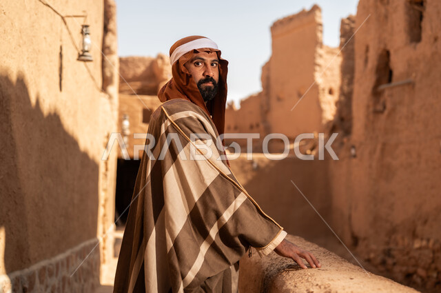 صورة من الجانب رجل عربي سعودي خليجي يرتدي زي يوم التأسيس يقف بجانب مبنى طيني, ذكرى تأسيس الدوله السعوديه الاولى على يد الامام محمد بن سعود, يوم بدينا 22 فبراير 1727م, العمق التاريخي والحضاري للمملكة، هوية اليوم الوطني السعودي 92