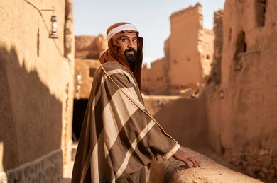 A side view of an Arab Saudi Gulf man wearing the Foundation Day attire standing next to a mud building, commemorating the establishment of the first Saudi state by Imam Muhammad bin Saud, which began on February 22, 1727. The historical and cultural depth of the Kingdom, the identity of Saudi National Day 92.