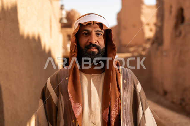 رجل كبير السن عربي سعودي خليجي يرتدي زي يوم التأسيس أمام مبنى طيني, ذكرى تأسيس الدوله السعوديه الاولى على يد الامام محمد بن سعود, يوم بدينا 22 فبراير 1727م, العمق التاريخي والحضاري للمملكة، ارتداء الأزياء التقليدية تعكس التراث