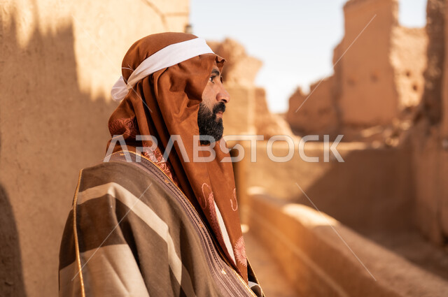صورة من الجانب لرجل كبير السن عربي سعودي خليجي يرتدي زي يوم التأسيس داخل مبنى طيني, ذكرى تأسيس الدوله السعوديه الاولى على يد الامام محمد بن سعود, يوم بدينا 22 فبراير 1727م, العمق التاريخي والحضاري للمملكة، ارتداء الأزياء التقليدية تعكس التراث