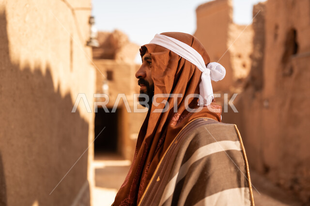 صورة من الجانب لرجل كبير السن عربي سعودي خليجي يرتدي زي يوم التأسيس داخل مبنى طيني, ذكرى تأسيس الدوله السعوديه الاولى على يد الامام محمد بن سعود, يوم بدينا 22 فبراير 1727م, العمق التاريخي والحضاري للمملكة، ارتداء الأزياء التقليدية تعكس التراث