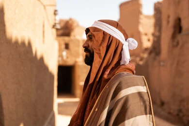 A side view of an elderly Arab Saudi Gulf man wearing the Foundation Day attire inside a mud building, commemorating the establishment of the first Saudi state by Imam Muhammad bin Saud, on the day we began, February 22, 1727 AD, the historical and cultural depth of the Kingdom, wearing traditional clothing reflects the heritage.