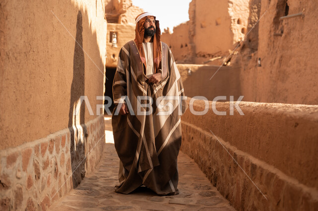 رجل كبير السن عربي سعودي خليجي يرتدي زي يوم التأسيس أمام مبنى طيني, ذكرى تأسيس الدوله السعوديه الاولى على يد الامام محمد بن سعود, يوم بدينا 22 فبراير 1727م, العمق التاريخي والحضاري للمملكة، ارتداء الأزياء التقليدية تعكس التراث