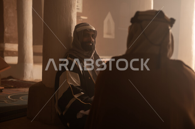 رجل كبير السن عربي سعودي خليجي يرتدي البشت وعقال يوم التأسيس يجلس مع رجل داخل مبنى طيني, ذكرى تأسيس الدوله السعوديه الاولى على يد الامام محمد بن سعود, يوم بدينا 22 فبراير 1727م, العمق التاريخي والحضاري للمملكة، ارتداء الأزياء التقليدية تعكس التراث