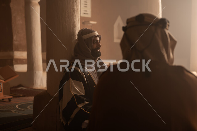 رجل كبير السن عربي سعودي خليجي يرتدي البشت وعقال يوم التأسيس يجلس مع رجل داخل مبنى طيني, ذكرى تأسيس الدوله السعوديه الاولى على يد الامام محمد بن سعود, يوم بدينا 22 فبراير 1727م, العمق التاريخي والحضاري للمملكة، ارتداء الأزياء التقليدية تعكس التراث
