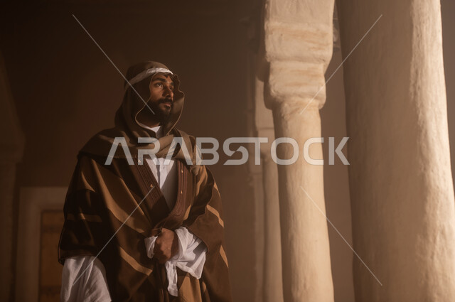 رجل كبير السن عربي سعودي خليجي يرتدي البشت وعقال يوم التأسيس يقف  داخل مبنى طيني تفاصيل العمارة القديمة, ذكرى تأسيس الدوله السعوديه الاولى على يد الامام محمد بن سعود, يوم بدينا 22 فبراير 1727م, العمق التاريخي والحضاري للمملكة، ارتداء الأزياء التقليدية تعكس التراث