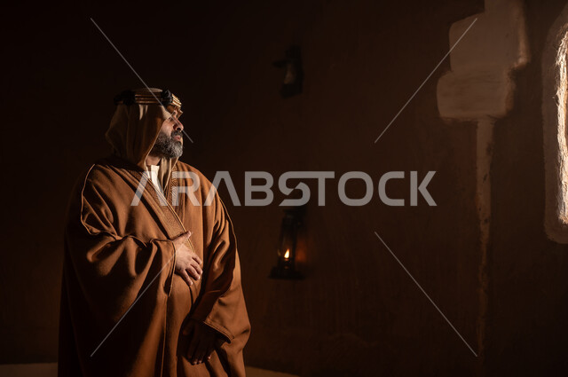رجل كبير السن عربي سعودي خليجي يرتدي البشت وعقال يوم التأسيسيقف يجانب شباك داخل مبنى طيني , ذكرى تأسيس الدوله السعوديه الاولى على يد الامام محمد بن سعود, يوم بدينا 22 فبراير 1727م, العمق التاريخي والحضاري للمملكة، ارتداء الأزياء التقليدية تعكس التراث