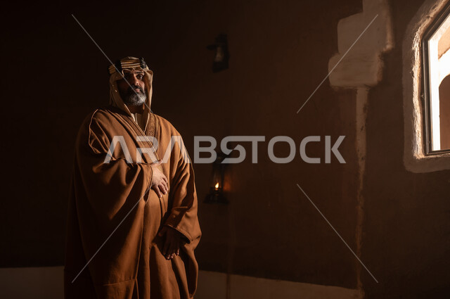 رجل كبير السن عربي سعودي خليجي يرتدي البشت وعقال يوم التأسيسيقف يجانب شباك داخل مبنى طيني , ذكرى تأسيس الدوله السعوديه الاولى على يد الامام محمد بن سعود, يوم بدينا 22 فبراير 1727م, العمق التاريخي والحضاري للمملكة، ارتداء الأزياء التقليدية تعكس التراث