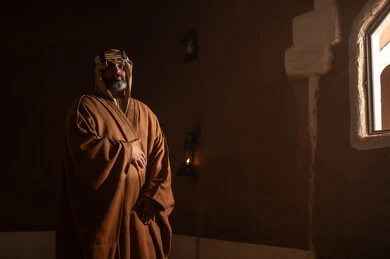 An elderly Arab Saudi Gulf man wearing a bisht and agal stands next to a window inside a mud building, commemorating the founding of the first Saudi state by Imam Muhammad bin Saud, on the day we began, February 22, 1727 AD. The historical and cultural depth of the Kingdom, wearing traditional attire reflects the heritage.
