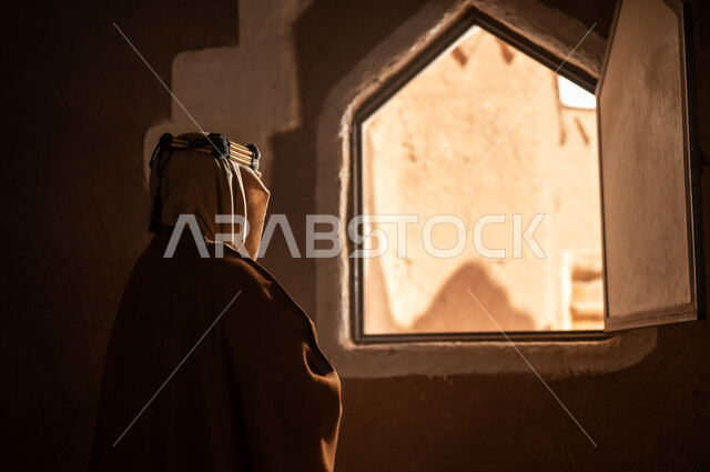 صورة من الخلف لرجل كبير السن عربي سعودي خليجي يرتدي البشت وعقال يوم التأسيسيقف يجانب شباك داخل مبنى طيني , ذكرى تأسيس الدوله السعوديه الاولى على يد الامام محمد بن سعود, يوم بدينا 22 فبراير 1727م, العمق التاريخي والحضاري للمملكة، ارتداء الأزياء التقليدية تعكس التراث