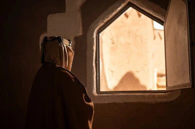 صورة من الخلف لرجل كبير السن عربي سعودي خليجي يرتدي البشت وعقال يوم التأسيسيقف يجانب شباك داخل مبنى طيني , ذكرى تأسيس الدوله السعوديه الاولى على يد الامام محمد بن سعود, يوم بدينا 22 فبراير 1727م, العمق التاريخي والحضاري للمملكة، ارتداء الأزياء التقليدية تعكس التراث