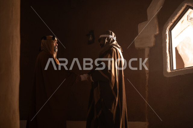رجل كبير السن عربي سعودي خليجي يرتدي البشت وعقال يوم التأسيس يتحدث داخل مبنى طيني مع رجل, ذكرى تأسيس الدوله السعوديه الاولى على يد الامام محمد بن سعود, يوم بدينا 22 فبراير 1727م, العمق التاريخي والحضاري للمملكة، ارتداء الأزياء التقليدية تعكس التراث