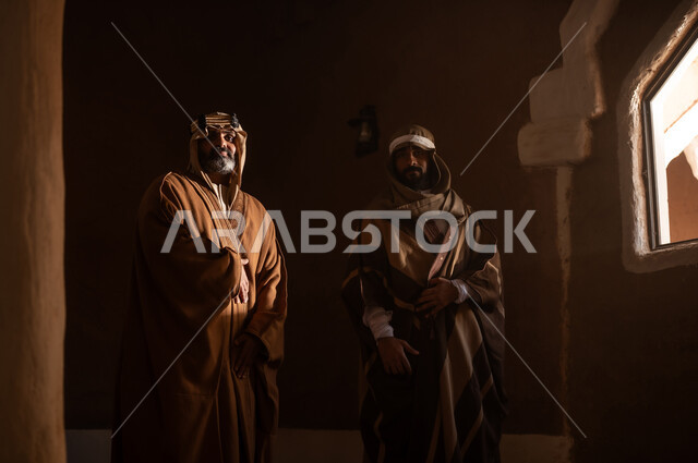 رجل كبير السن عربي سعودي خليجي يرتدي البشت وعقال يوم التأسيس يقف داخل مبنى طيني مع رجل اخر, ذكرى تأسيس الدوله السعوديه الاولى على يد الامام محمد بن سعود, يوم بدينا 22 فبراير 1727م, العمق التاريخي والحضاري للمملكة، ارتداء الأزياء التقليدية تعكس التراث