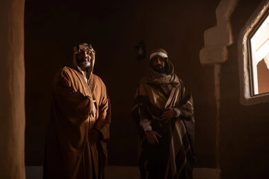 An elderly Arab Saudi Gulf man wearing a bisht and agal on Foundation Day stands inside a mud building with another man, commemorating the establishment of the first Saudi state by Imam Muhammad bin Saud, on the day we began, February 22, 1727 AD. The historical and cultural depth of the Kingdom, wearing traditional attire reflects the heritage.
