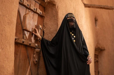 Wearing traditional dress on the anniversary of the founding of the first Saudi state, February 22nd, expresses pride in national identity and reflects the Bedouin lifestyle in traditional mud houses. The day we began, 1727 AD, a Saudi Arabian woman from the Gulf region wearing a black abaya and burqa in an old house.