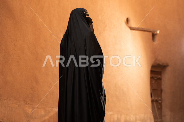 A side view of a Saudi Arabian woman wearing a black abaya and burqa in an old house, wearing traditional dress on the anniversary of the founding of the first Saudi state, February 22nd, expressing pride in national identity and the traditional Bedouin lifestyle in heritage mud houses, the day we began, 1727 AD