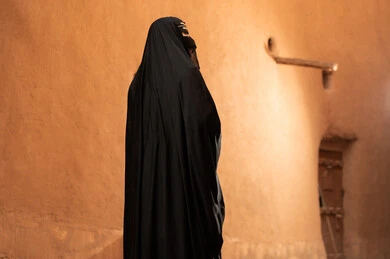 صورة من الجانب لامرأة عربية خليجية سعودية ترتدي العباءة السوداء والبرقع في منزل قديم، ارتداء الزي الشعبي في ذكرى تأسيس الدولة السعودية الاولى 22 فبراير، الفخر والاعتزاز بالهوية الوطنية، نمط حياة البدو الشعبية في البيوت الطينية التراثية، يوم بدينا 1727 م