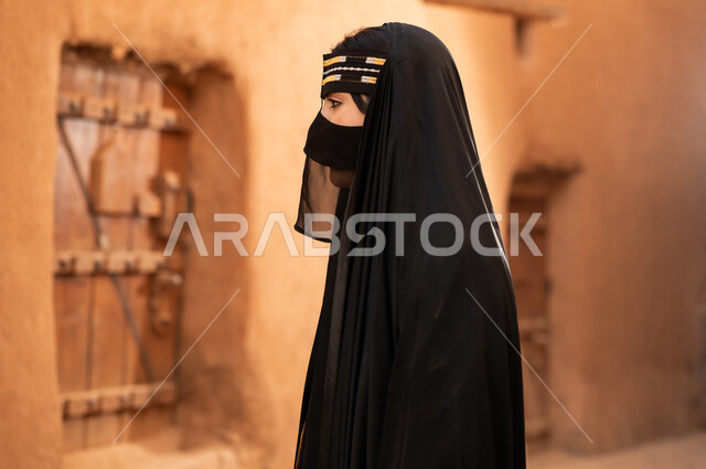 A side view of a Saudi Arabian woman wearing a black abaya and burqa in an old house, wearing traditional dress on the anniversary of the founding of the first Saudi state, February 22nd, expressing pride in national identity and the traditional Bedouin lifestyle in heritage mud houses, the day we began, 1727 AD