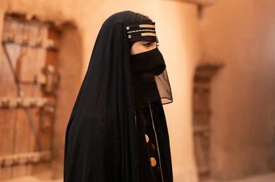 صورة من الجانب لامرأة عربية خليجية سعودية ترتدي العباءة السوداء والبرقع في منزل قديم، ارتداء الزي الشعبي في ذكرى تأسيس الدولة السعودية الاولى 22 فبراير، الفخر والاعتزاز بالهوية الوطنية، نمط حياة البدو الشعبية في البيوت الطينية التراثية، يوم بدينا 1727 م