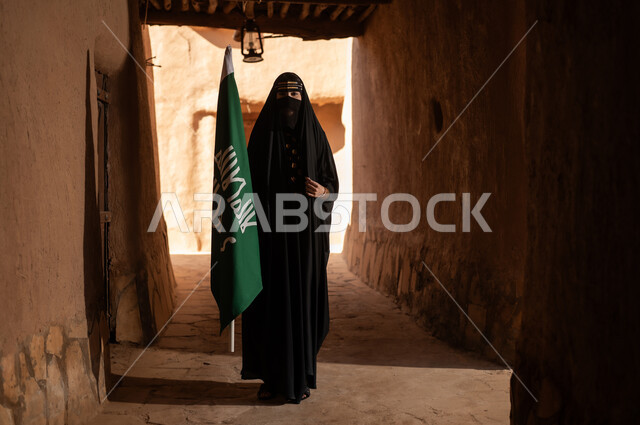 امرأة عربية خليجية سعودية ترتدي العباءة السوداء والبرقع في منزل قديم تمسك بيدها راية التأسيس، ارتداء الزي الشعبي في ذكرى تأسيس الدولة السعودية الاولى 22 فبراير، الفخر والاعتزاز بالهوية الوطنية، نمط حياة البدو الشعبية في البيوت الطينية التراثية، يوم بدينا 1727 م