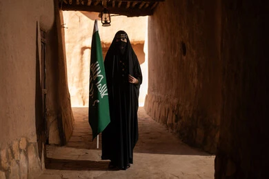 A Saudi Arabian woman from the Gulf region, wearing a black abaya and burqa, holds the flag of the founding of the first Saudi state in an old house. She wears traditional dress on the anniversary of the founding of the first Saudi state, February 22nd, expressing pride in national identity and reflecting the Bedouin lifestyle in traditional mud houses. The day we began, 1727 AD.