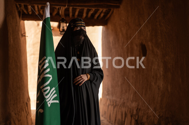 On the day we began, 1727 AD, a Saudi Arabian Gulf woman wearing a black abaya and burqa in an old house, holding the flag of the founding, wearing traditional dress on the anniversary of the founding of the first Saudi state, February 22, pride and honor in national identity, the popular Bedouin lifestyle in traditional mud houses.