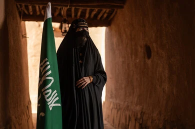 يوم بدينا 1727 م، امرأة عربية خليجية سعودية ترتدي العباءة السوداء والبرقع في منزل قديم تمسك بيدها راية التأسيس، ارتداء الزي الشعبي في ذكرى تأسيس الدولة السعودية الاولى 22 فبراير، الفخر والاعتزاز بالهوية الوطنية، نمط حياة البدو الشعبية في البيوت الطينية التراثية