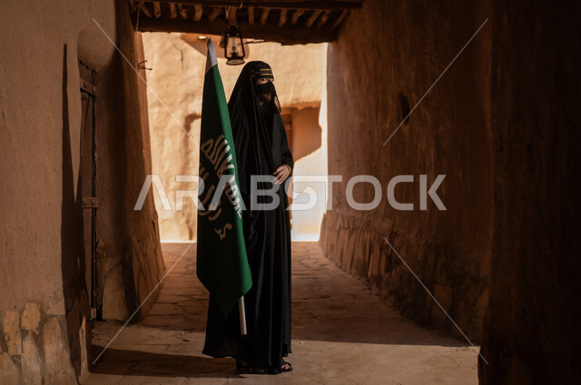 امرأة عربية خليجية سعودية ترتدي العباءة السوداء والبرقع في منزل قديم تمسك بيدها راية التأسيس، ارتداء الزي الشعبي في ذكرى تأسيس الدولة السعودية الاولى 22 فبراير، الفخر والاعتزاز بالهوية الوطنية، نمط حياة البدو الشعبية في البيوت الطينية التراثية، يوم بدينا 1727 م